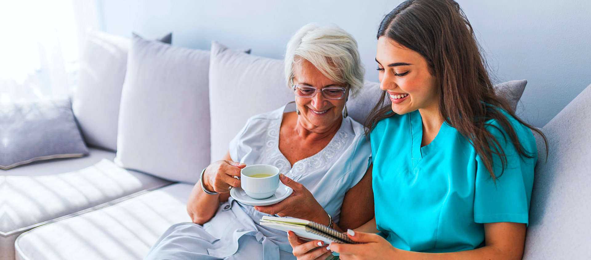 caregiver reading the book and senior woman are listening while drinking a cup of tea